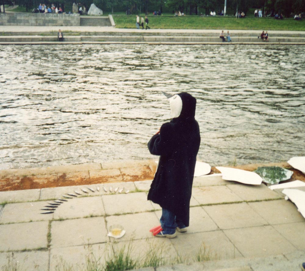 Back view of a child in a penguin costume facing the river with fish arranged on the ground