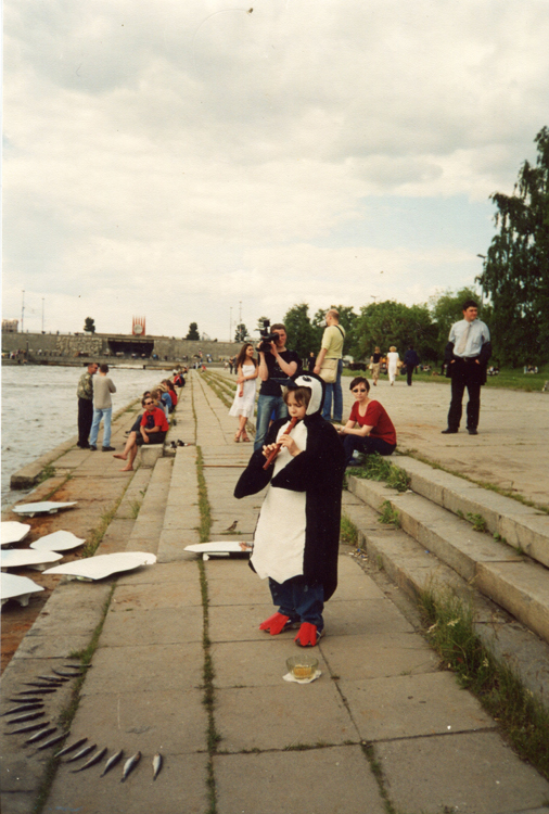 Child in a penguin costume playing a wooden flute with fish arranged in a semicircle on a river embankment